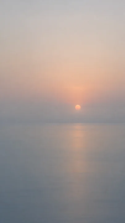 一轮淡杏色初阳浮在海雾里，柔和暖光落向灰蓝海面，朦胧海天留出安静的锁屏空间。，手机壁纸